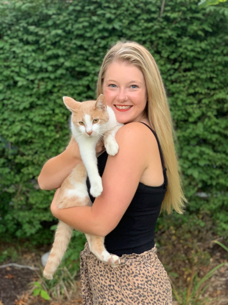 Blonde girl in black tank top holding an orange and white cat while standing in front of a green bush