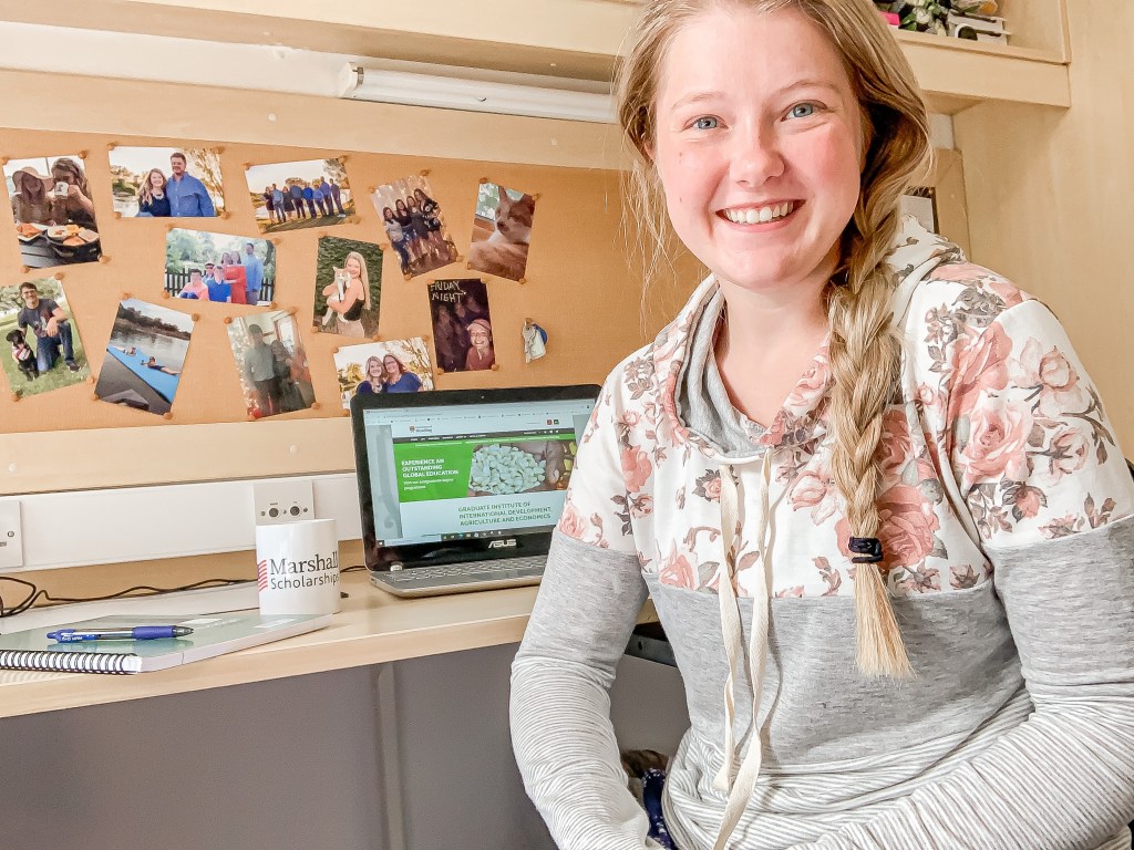 A blonde girl wearing a flowery hoodie while sitting in front of a desk with a laptop, coffee mug, notebook, and pen sitting on it and a bulletin board with pictures pinned on it in the background
